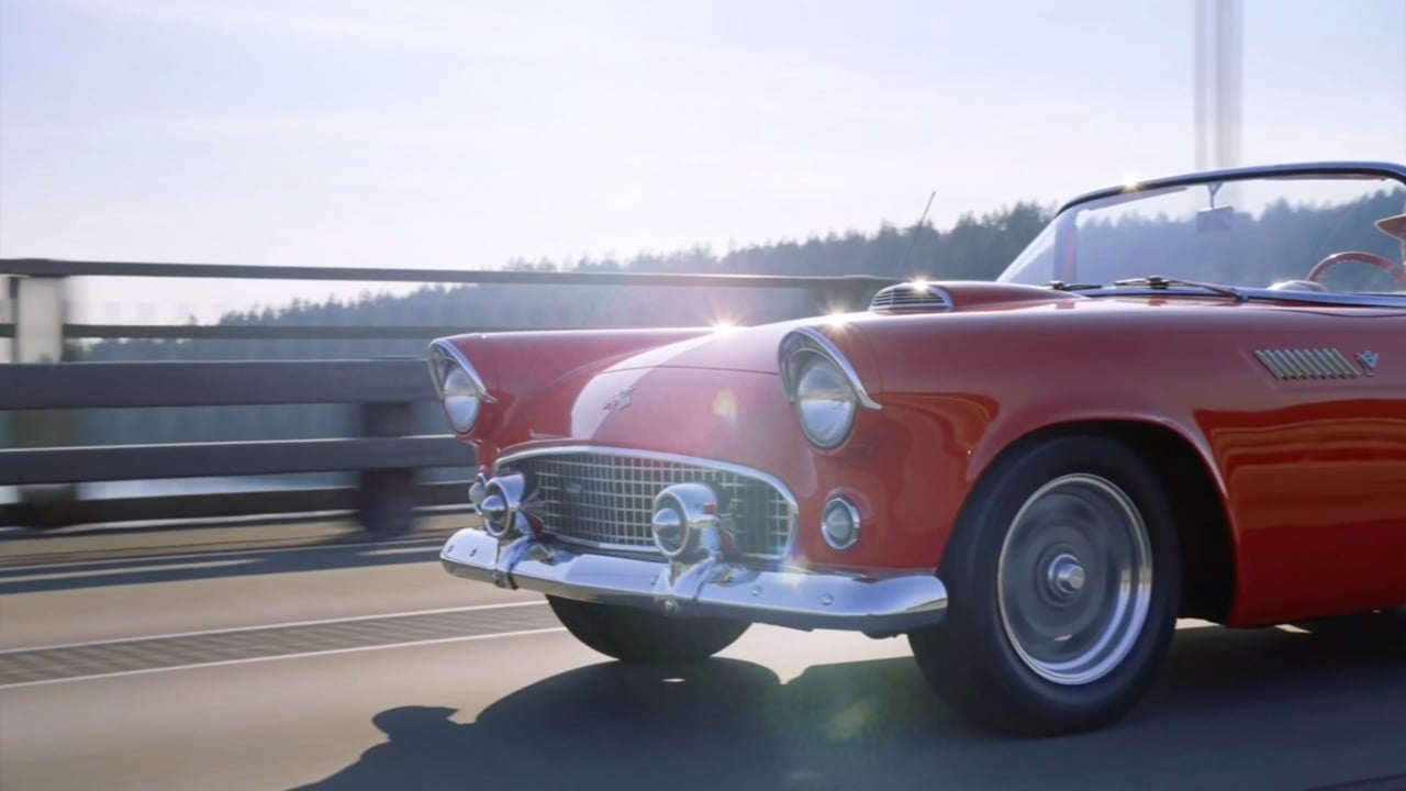 The cherry red 1955 Ford Thunderbird driving across a bridge in low sunlight, chrome grille and headlights gleaming.